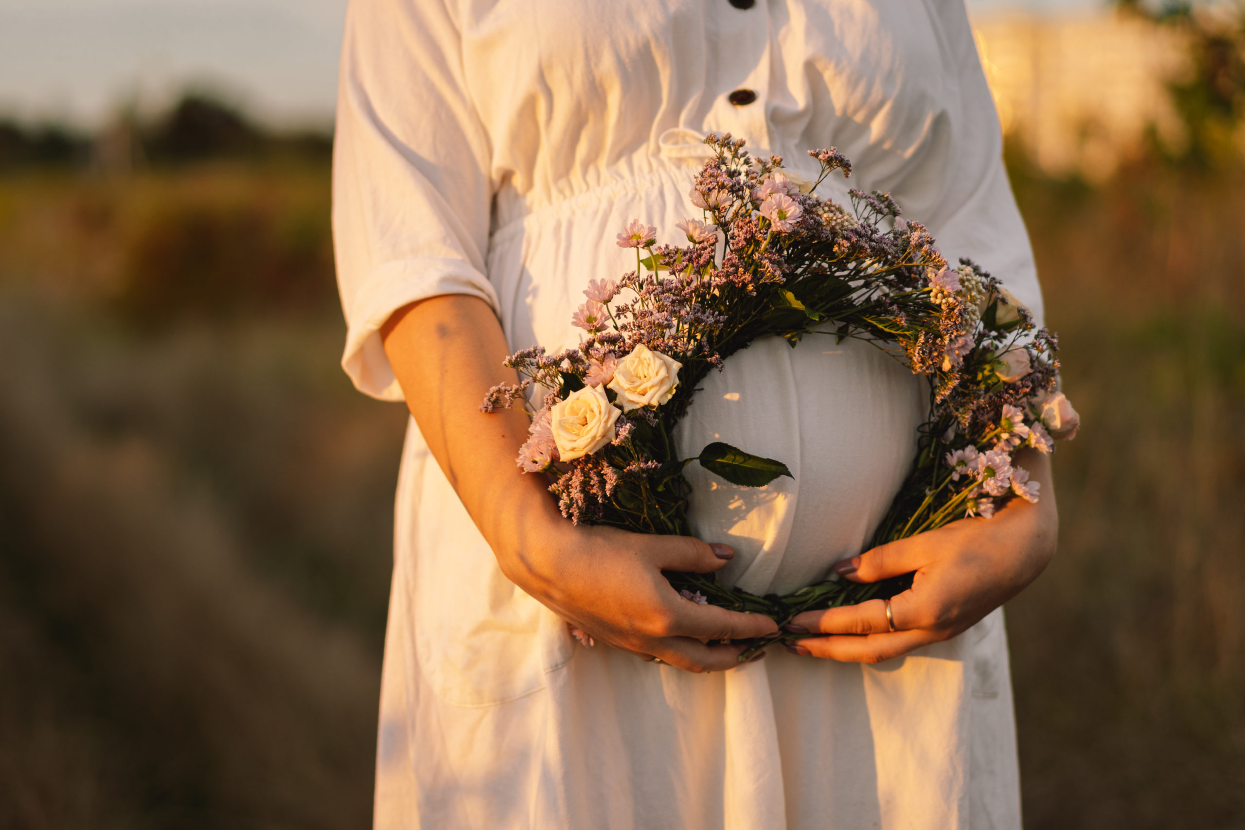 Schwangere Frau steht in weißem Leinenkleid, mit halblangem Arm, auf einem Feld und hält einen üppig gebundenen Blumenkranz, vor ihrem Babybauch.