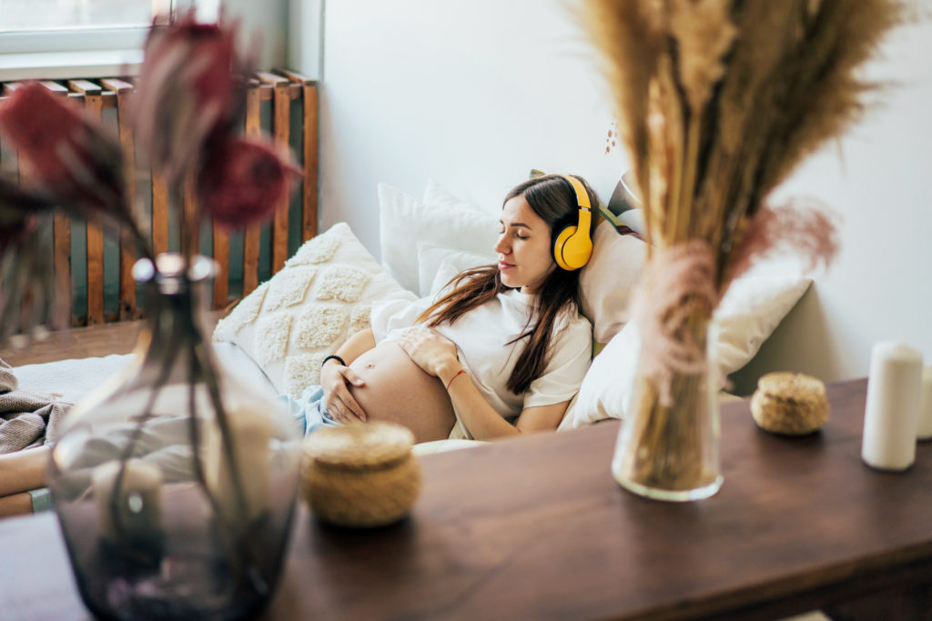 Schwangere Frau mit langen braunen Haaren und gelben Kopfhörern liegt mit geschlossenen Augen entspannt auf Ihrem Bett. Sie hört eine geburtsvorbereitende Meditation.