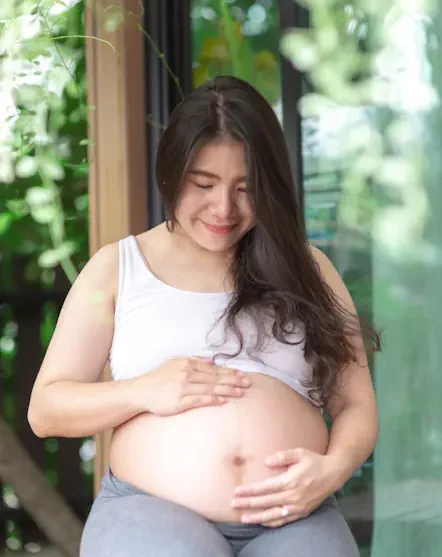 Pränatale Bindung: Schwangere Frau, auf einem Balkon im Grünen. Ihre Hände liegen auf ihrem nackten Bauch. Mit liebevollen Berührungen geht sie in Kontakt mit ihrem Ungeborenen.